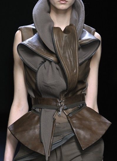 a female model wearing a fashionable leather vest with multiple zippers. the vest has a high collar and is styled with a skirt and a belt. the model is standing on a runway with a dark background.