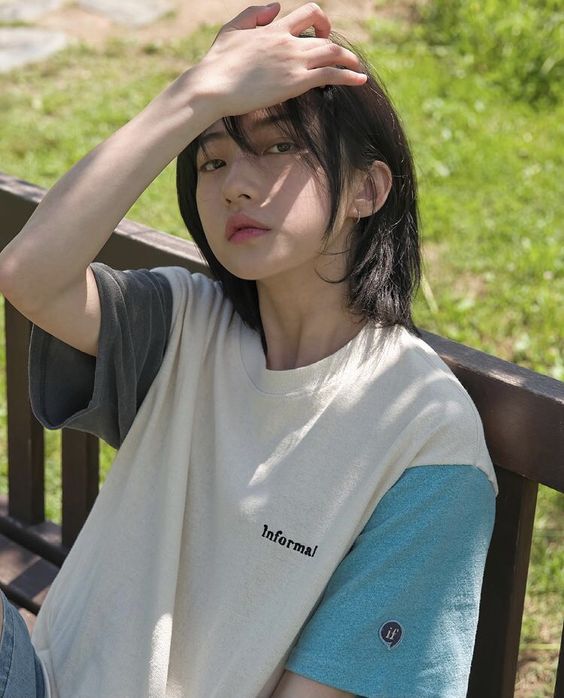 a young woman with short dark hair and wearing a twotoned tshirt is sitting on a wooden bench outdoors. she is looking directly at the camera with her left hand resting on her head. the sunlight is dappled, creating a mix of light and shadow on her face and the background. the overall mood is casual and relaxed.