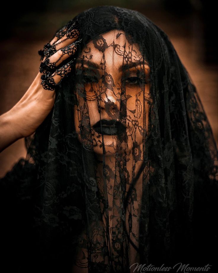 the image is a closeup portrait of a woman partially obscured by a black lace veil. the veil is draped over her head and covers most of her face, only revealing her eyes and the outline of her lips. she's wearing dark makeup and black lace gloves. the lighting is dramatic, casting shadows and highlighting the texture of the lace. the background is dark and out of focus, drawing attention to the model. the overall mood is mysterious, sensual, and gothic. the watermark "motionless moments" suggests the photographer's name or brand.