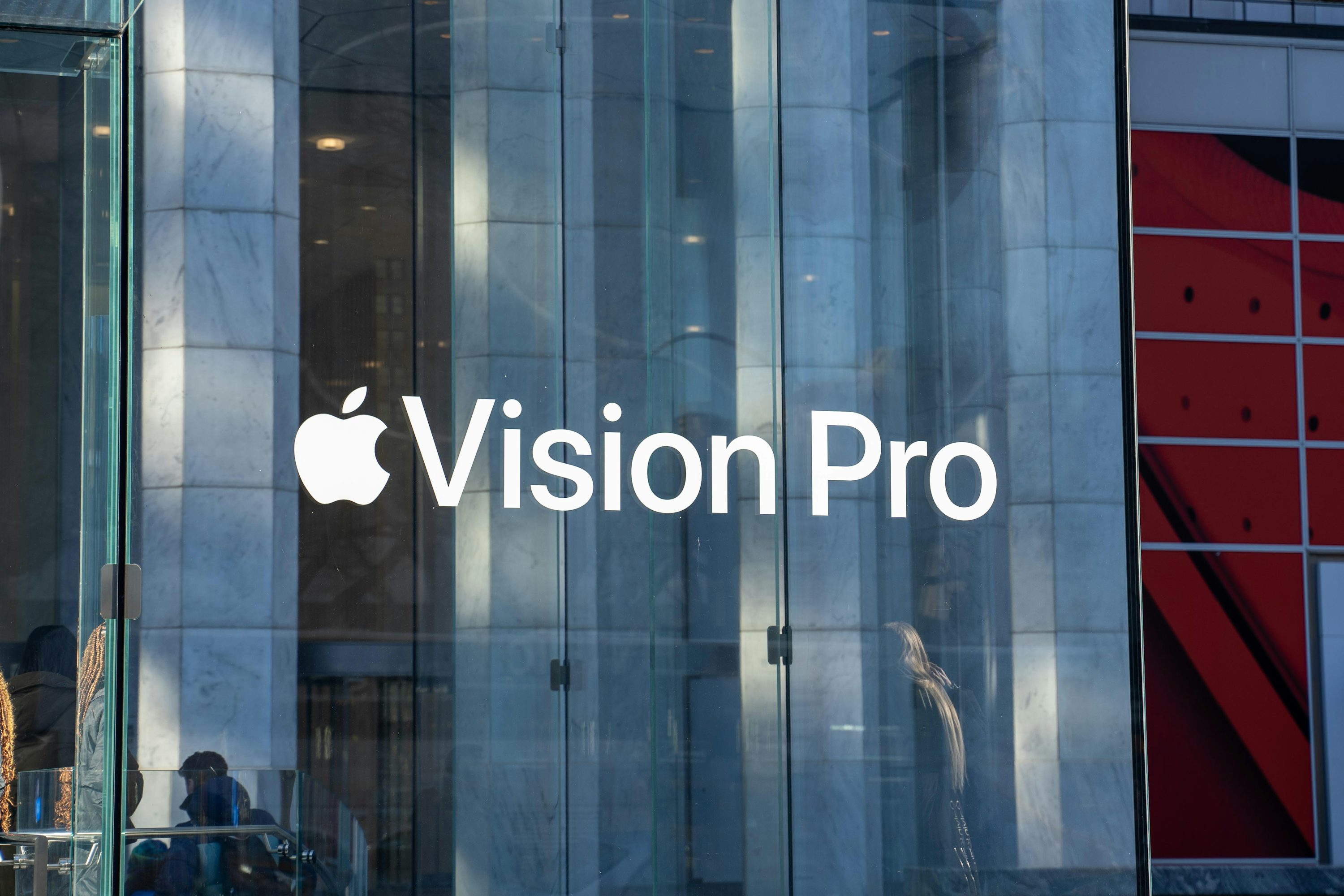 the image shows the apple vision pro logo on the glass storefront of an apple store. the reflection on the glass shows the interior of the store and the architecture of the building.