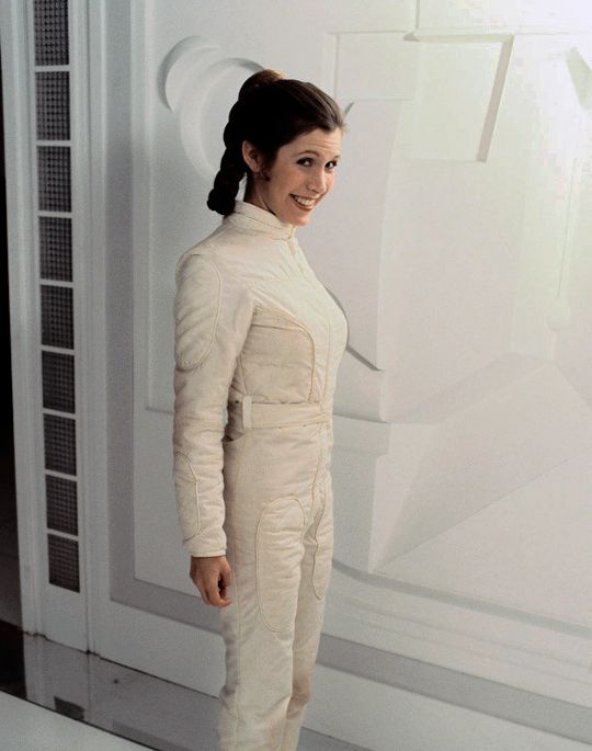 a portrait of carrie fisher as princess leia from star wars in her iconic white jumpsuit. she is smiling and looking at the camera.  she is standing in a white room, and there is a hint of a white, textured wall in the background.  the image evokes a sense of classic science fiction and the iconic character of princess leia.