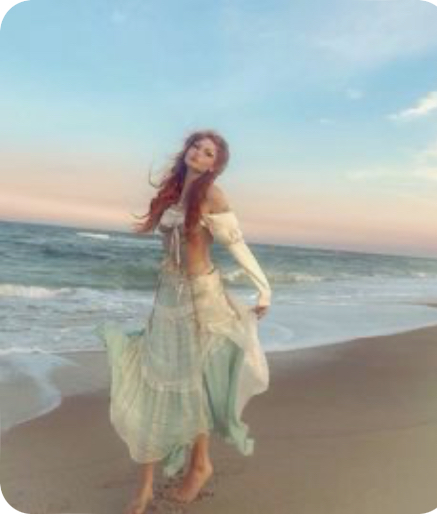 a redhaired woman in a flowing, pale green boho dress stands on a beach at sunset. the ocean is behind her and the setting sun casts a warm glow on the scene.
