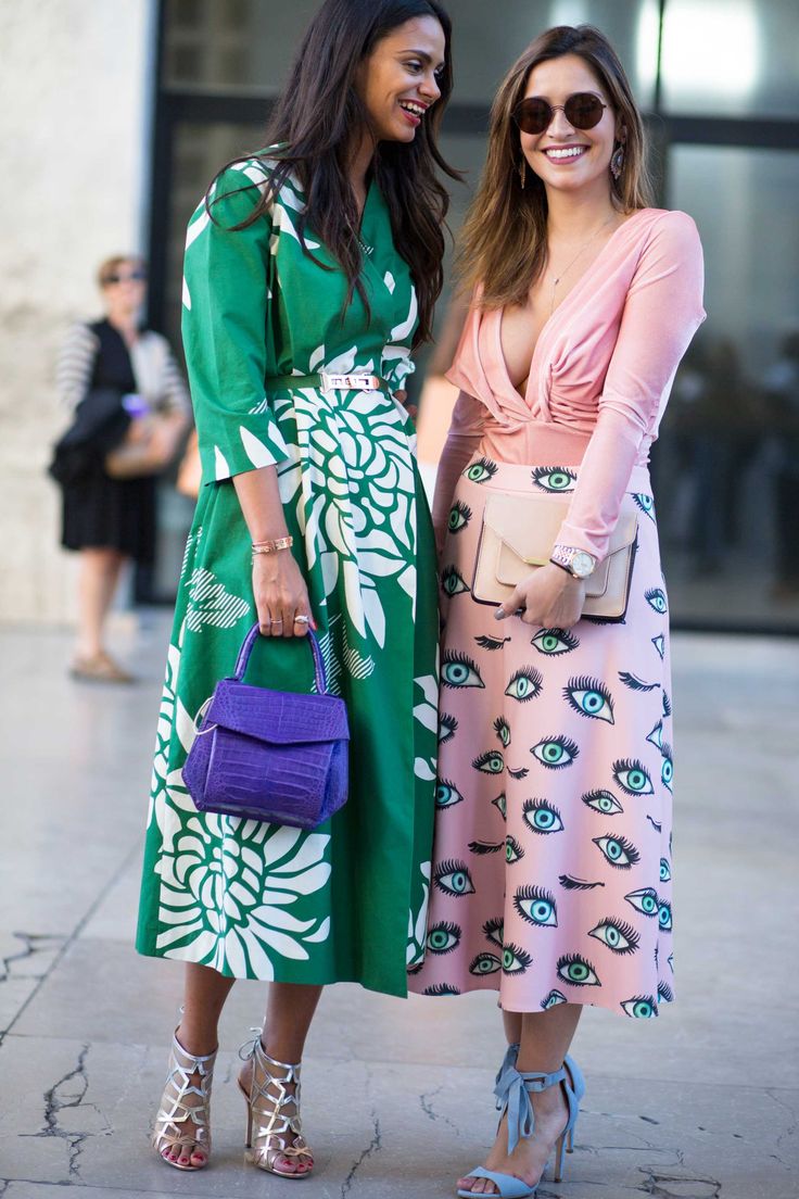 two stylishly dressed women are standing on a city sidewalk,  chatting and smiling. one woman wears a long, green floral dress paired with silver strappy heels and a purple handbag.  the other woman sports a pink, eyeprint midiskirt and a light pink longsleeved top, along with blue suede heels and a beige clutch. they both wear sunglasses and appear to be enjoying themselves during a fashion event or outing. the background is slightly blurred but shows a modern city setting.