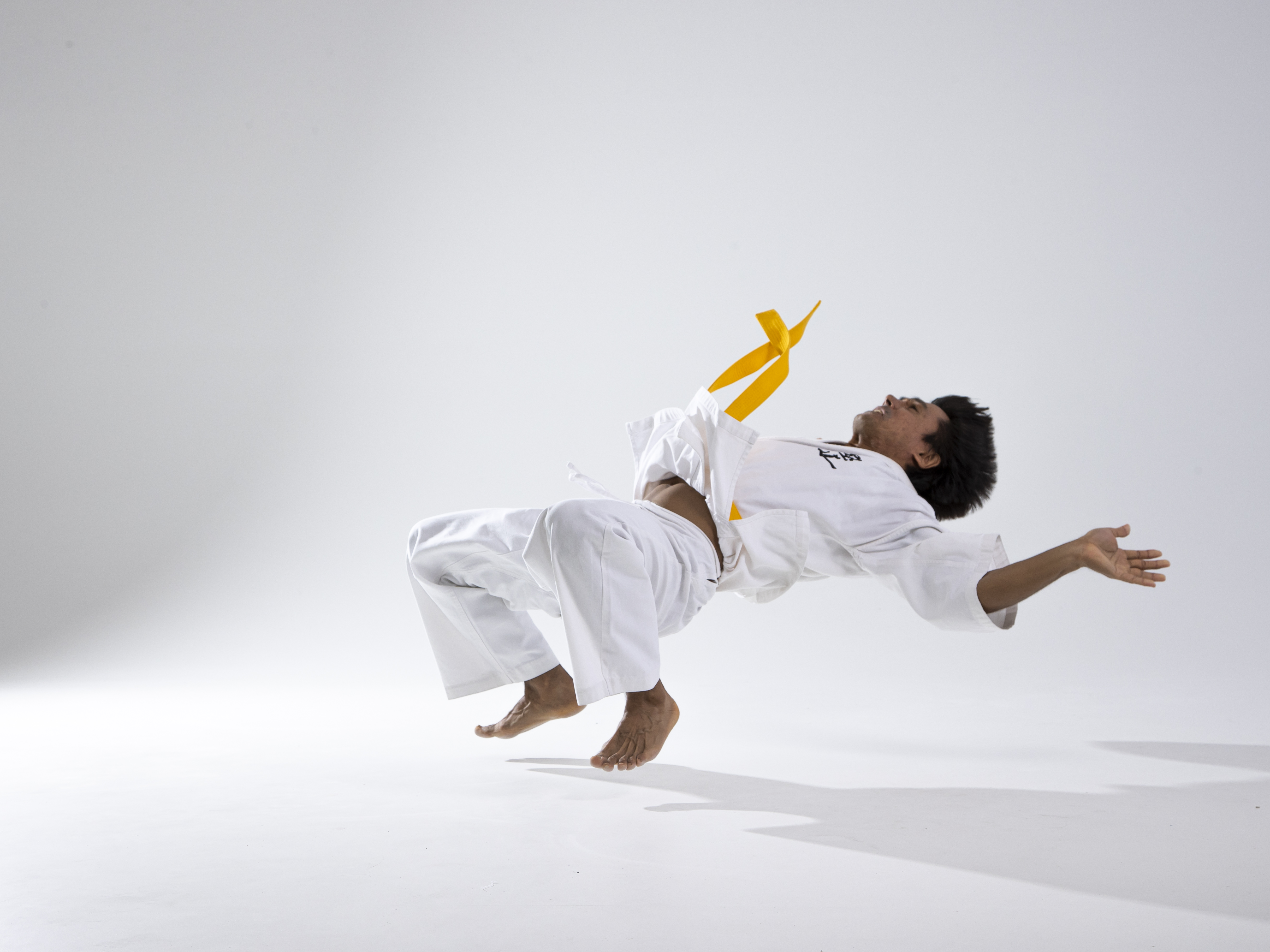 a man in a white karate or taekwondo gi with a yellow belt is performing a midair jump or kick in a studio setting. he is barefoot and appears to be in motion, with his body curved and his arms outstretched. the background is a plain white, and a shadow is cast beneath him.