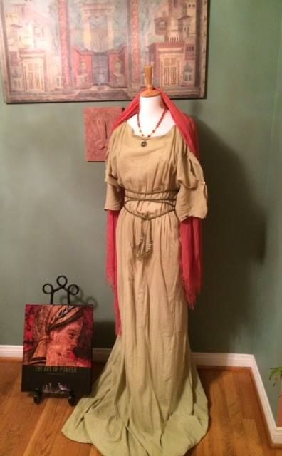 the image shows a floorlength, light beige romanstyle dress displayed on a mannequin against a wall. the dress is simple, with a loose fit and tied at the waist.  it's accessorized with a coralcolored shawl draped around the shoulders. the mannequin also wears a necklace.  a book titled "the art of pompeii" is visible on a stand in the foreground. an antiquestyle wall hanging featuring a roman fresco is displayed above the dress. the setting appears to be an interior with wooden flooring.  the overall style suggests a historical or theatrical costume display.