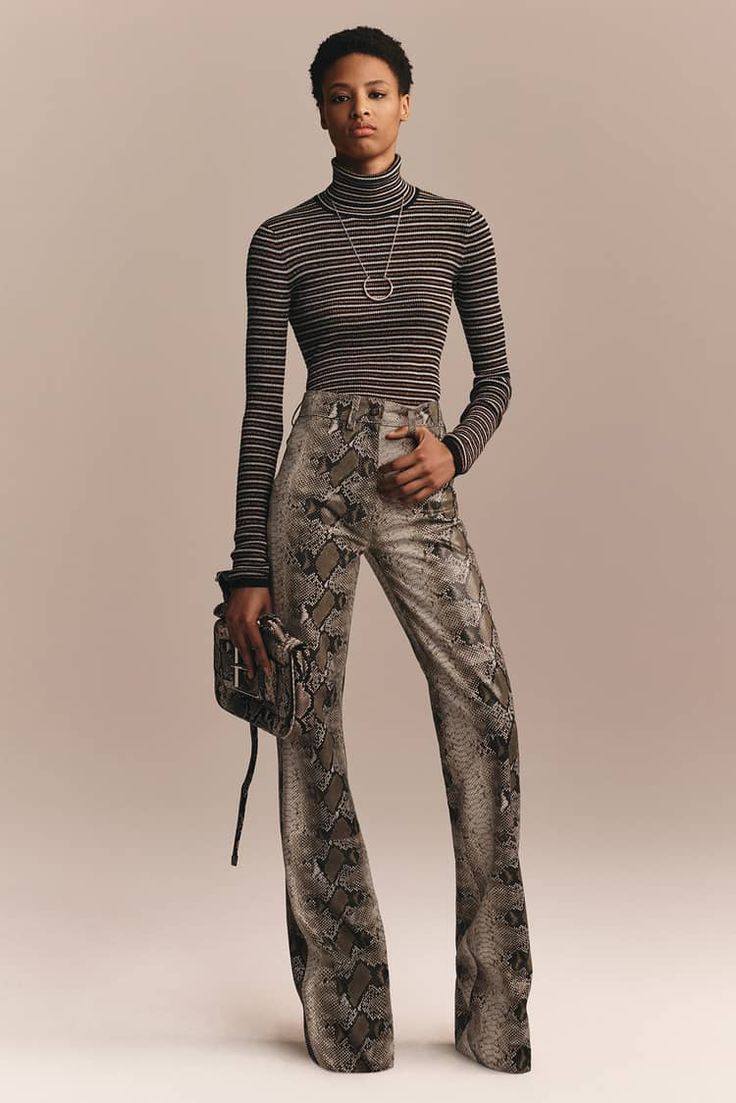 a young woman is posing in a studio setting. she is wearing a black and white striped turtleneck sweater, snakeskinprinted flared pants, and a small, black bag. she is also wearing a silver necklace.