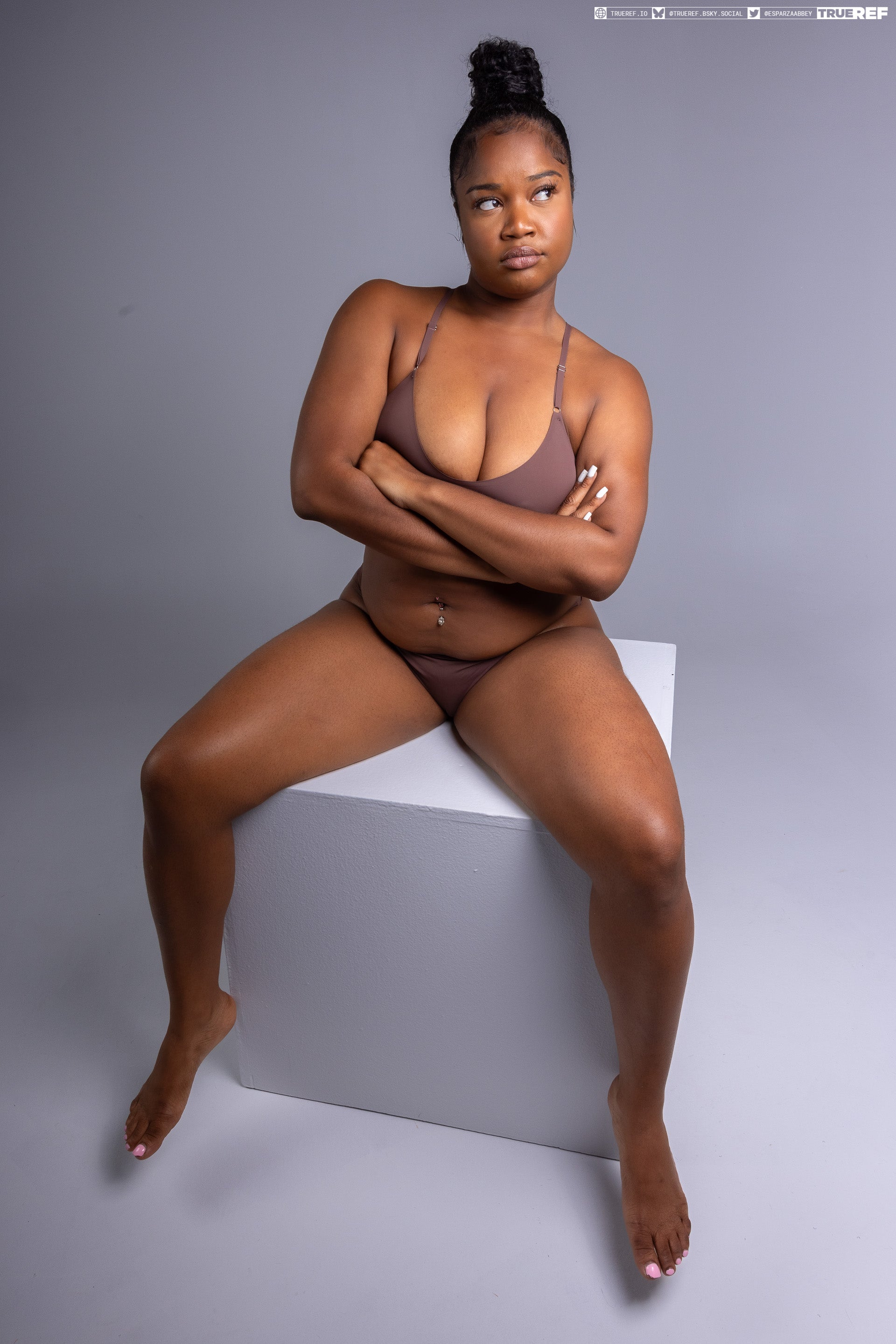 a darkskinned woman is sitting on a white cube, with her legs spread apart and arms crossed over her chest. she is wearing brown lingerie and has a serious expression on her face. the background is a solid gray color. the photo appears to be a studio shot. her hair is pulled up into a high bun.