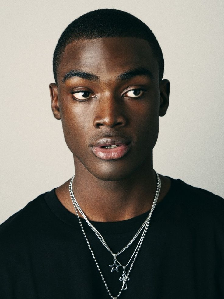 a young black man with short dark hair, wearing a black shirt and silver necklaces, looks down seriously. the image is a closeup portrait and shows his face and shoulders.