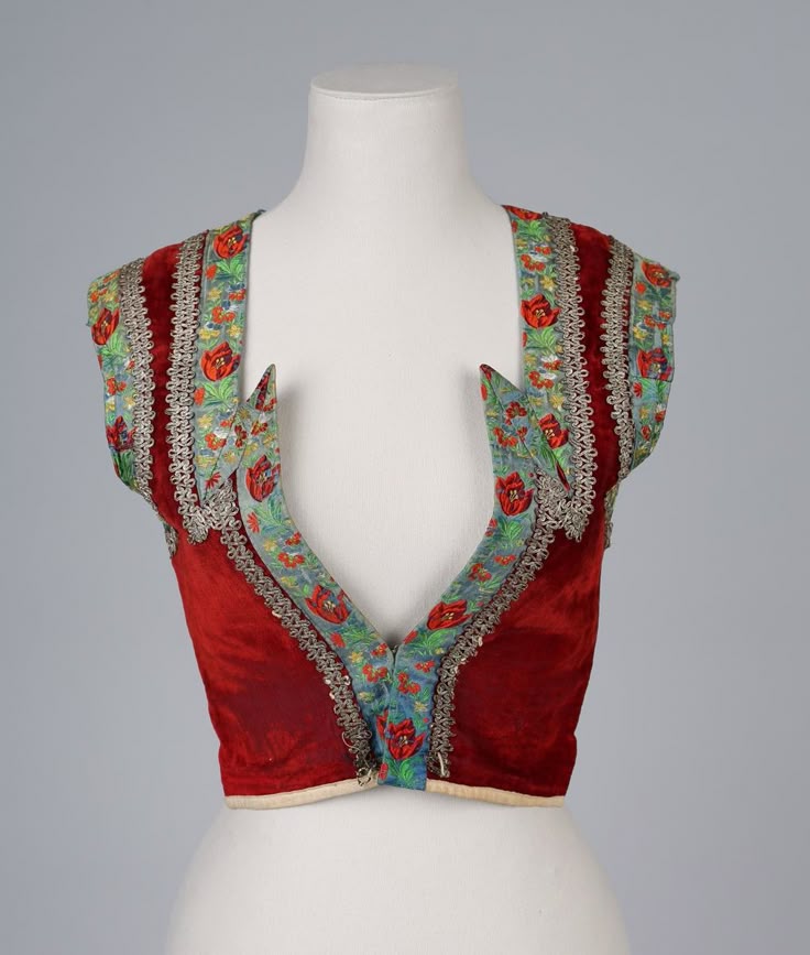 a sleeveless red velvet vest with blue floral embroidery and silver trim is displayed on a mannequin. the vest has a deep vneck and the floral pattern features red tulips. the silver trim borders the floral embroidery and edges of the vest.