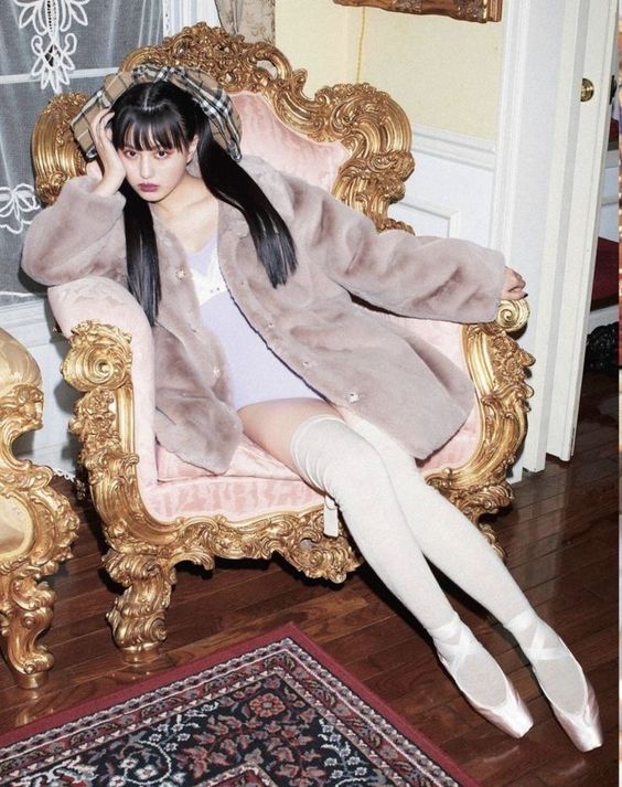 a young woman with long black hair and bangs is sitting on a pink and gold ornate chair. she is wearing a light purple dress, a long gray faux fur coat, white leg warmers, and pink ballet shoes. she is looking at the camera with a neutral expression. the room has a vintage feel, with floral wallpaper and a patterned rug.