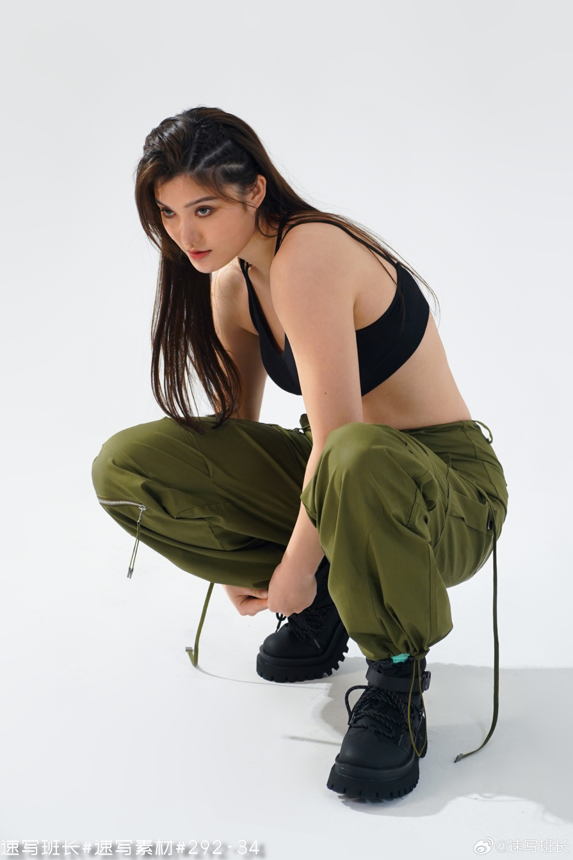 a young woman with long, dark hair styled with braids, is crouching in a studio. she is wearing a black bra top and olive green cargo pants with black, chunky boots. the image appears to be from a fashion photoshoot, with bright artificial lighting and an eyelevel perspective.