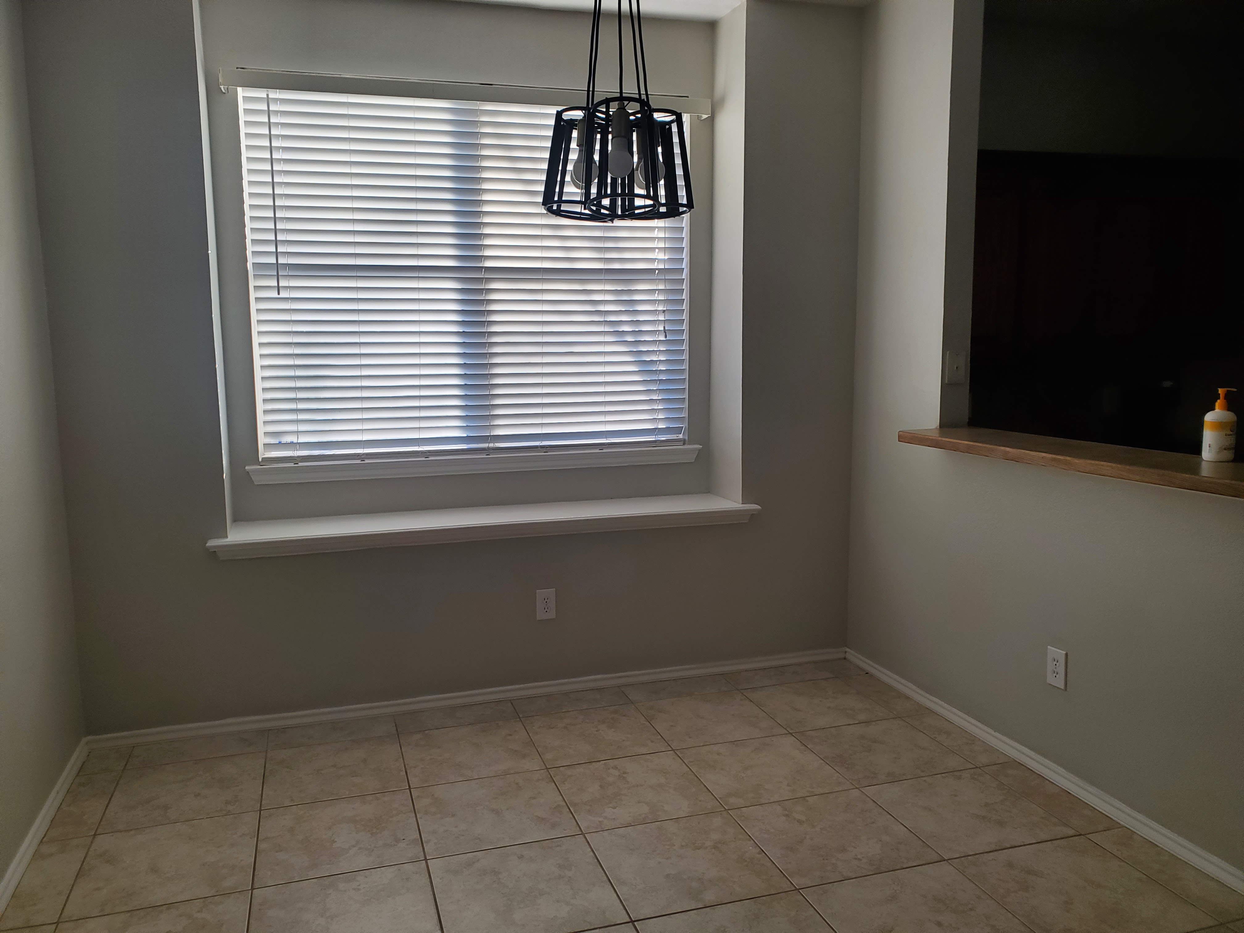 the image shows an interior room with a window covered by closed white blinds. a black geometric light fixture hangs in front of the window. the walls are painted a neutral grey color, and there is a tile floor. an electrical outlet is visible on one of the walls.