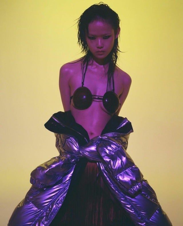 a woman with wet black hair and wearing a futuristic purple outfit with a silver jacket and a unique black bra is standing in a studio setting with yellow lighting. the woman is looking at the camera and has her hands on the jacket.