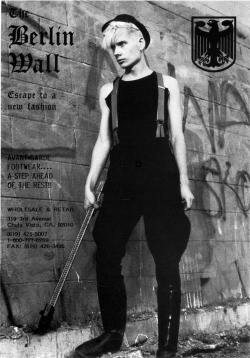 a black and white full shot of a young man, dressed in 1980sstyle punk or gothic fashion. he wears black pants and boots, suspenders, and a black top.  he has short bleached blonde hair and is wearing a newsboy cap. he stands against a graffiticovered brick wall with the words "the berlin wall" and advertising information overlaid on the image. the image has a gritty, urban feel. the overall aesthetic suggests a fashion advertisement for "avantgarde footwear." a german eagle emblem is also present on the wall.