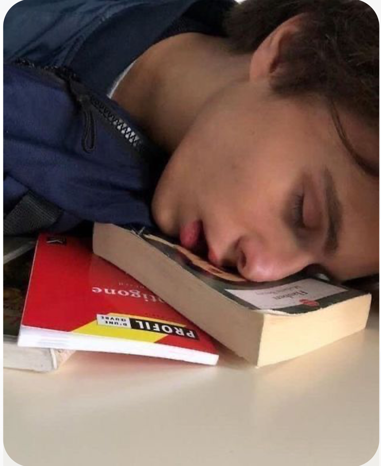a young man, presumably a student, is sleeping soundly on a desk with his face resting on a stack of books. the books are open, suggesting he was studying before falling asleep. the lighting is soft and natural, creating a peaceful atmosphere. the closeup perspective and the subject's relaxed posture emphasize his exhaustion.