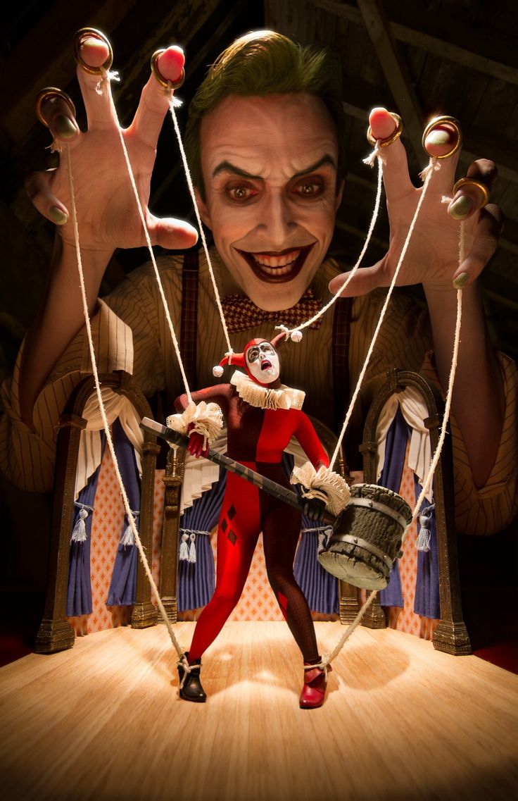 a man dressed as the joker is holding strings attached to a harley quinn puppet. joker is smiling menacingly while harley quinn is standing with a baseball bat. the scene is set up to appear like a puppet show.