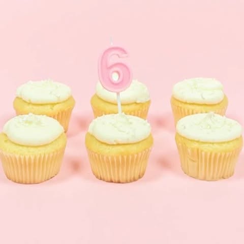 this image contains six cupcakes with white frosting on a pink background. one of the cupcakes has a pink birthday candle in the shape of the number six.