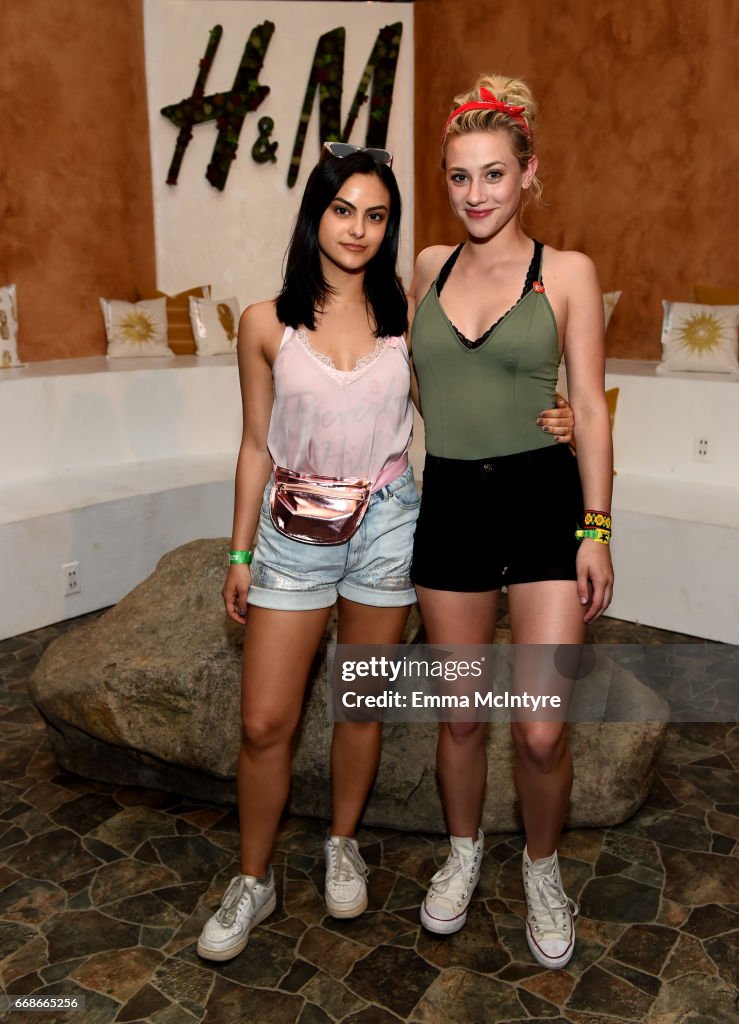 two women, lili reinhart and camila mendes, pose in front of an h&m sign. lili is wearing a green top and black shorts with white sneakers and a red headband. camila is wearing a pink tank top and denim shorts with white sneakers and sunglasses on her head. there is a large stone behind them and a tile floor.