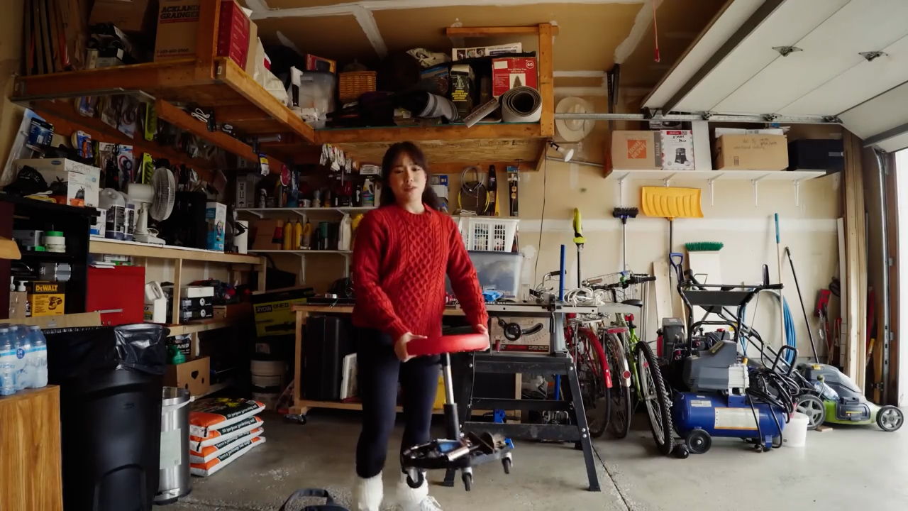 a woman wearing a red sweater and black pants stands in a cluttered garage. she is holding a red rolling stool and appears to be in the process of organizing or working on a project. the garage is filled with shelves overflowing with tools, equipment, and various items. there is a workbench in the background and bicycles leaning against the wall. the overall impression is one of a busy and wellused workspace.