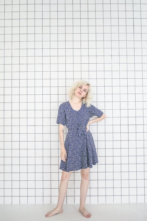 a woman with short blonde hair stands barefoot against a white tiled wall. she is wearing a kneelength dress with a blue floral pattern. the dress has short sleeves and a tie at the waist. she is in a studio setting.