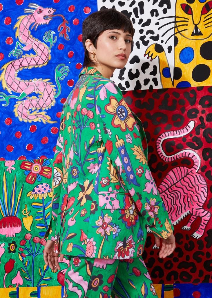the image showcases a young woman with a short, dark haircut modeling a vibrant green suit. the suit features a bold, eyecatching floral print with a variety of colors and whimsical designs.  the model is posed against a backdrop composed of a collage of colorful, handpainted illustrations featuring a dragon, tigers, and leopard patterns. the overall aesthetic is highly eclectic and artistic, showcasing both the fashion and the artwork in a striking contrast and complement. the bright colors and playful designs create a lively and fun mood.
