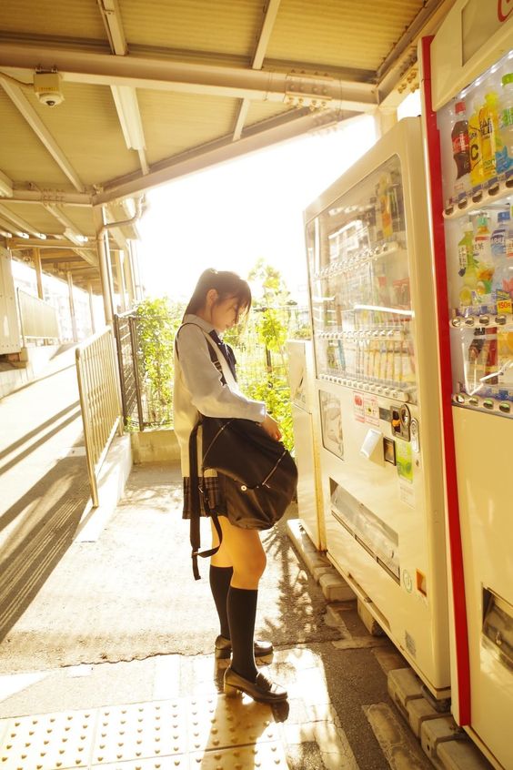 school girl in a blue and white uniform stands at a vending machine in a train station bathed in warm afternoon sunlight.  she is wearing black tights and shoes, and carries a large black shoulder bag. the sun is behind her, creating a backlit effect.