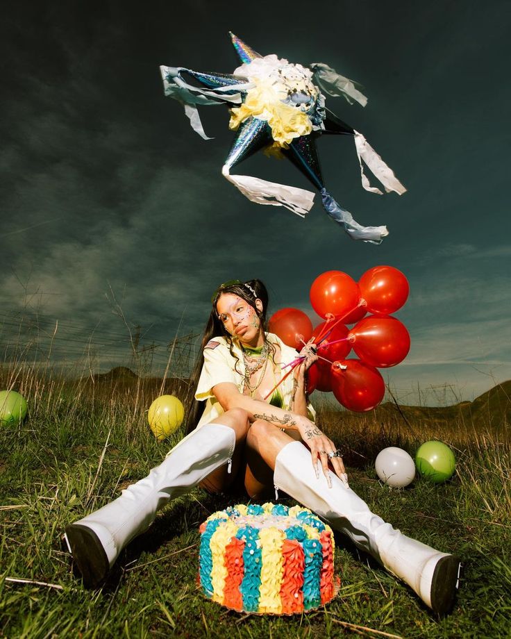 a young woman with dark hair and colorful face paint sits in a grassy field, holding a bunch of red balloons. she's wearing a yellow shortsleeved shirt, white thighhigh boots, and a necklace.  in front of her is a vibrant, colorful striped cake.  above her, a piñata hangs in the air against a dramatic sky.  other colorful balloons and a small cake are scattered around her. the overall scene is bright, festive, and suggestive of a birthday celebration.