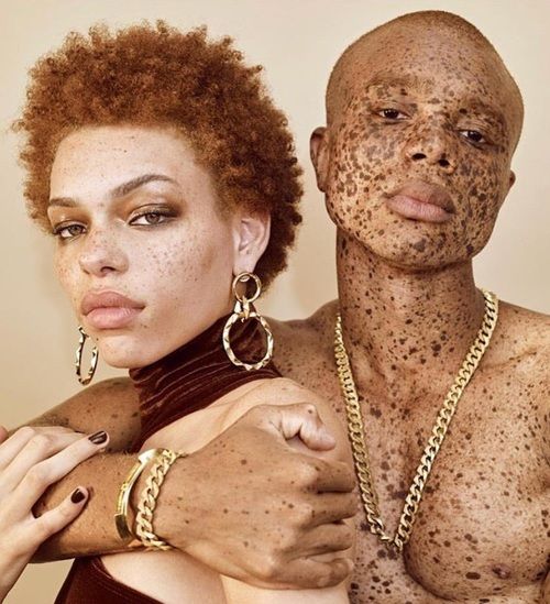 a closeup portrait of a couple embracing each other. the woman has red hair and freckles and is wearing gold earrings and a bracelet. the man has dark skin with freckles and is wearing a gold necklace. the couple is looking at each other with love and affection.