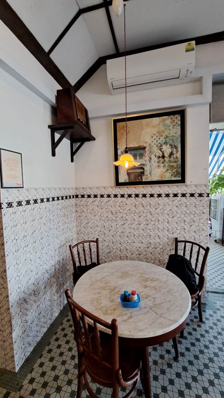 a small dining room with two chairs and a round table. the table is made of marble and is surrounded by two wooden chairs. the walls are covered in white tile with a black and white checkered pattern. there is a painting on the wall, a lamp hanging from the ceiling, and a wooden shelf with a brown wooden box. the floor is a checkered pattern of black and white tiles. the room has a vintage and classic feel, with simple and minimalist decor.