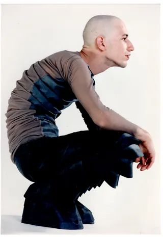 a bald man is sitting with his legs folded under him. he is wearing a light gray longsleeved shirt and black pants. the man is looking to the right, his right arm is bent, and his left arm is resting on his leg. he is wearing a small black watch. the background is a white wall.
