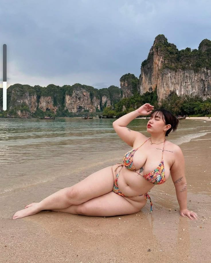 a woman is laying on a sandy beach wearing a bikini. the ocean water laps at the shore around her. she has short dark hair and tattoos. behind her are mountains and cliffs. the sky is cloudy.