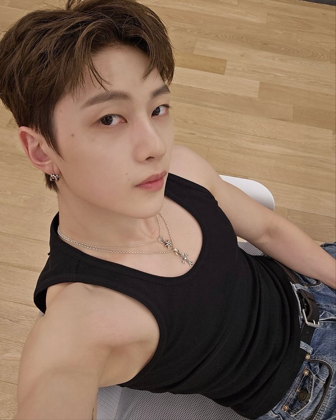 a young asian man with brown hair takes a selfie from a high angle. he is wearing a black tank top, blue jeans, and two silver chain necklaces. the photo appears to be taken indoors in a studio setting with bright, artificial lighting.  the floor is light brown wood.