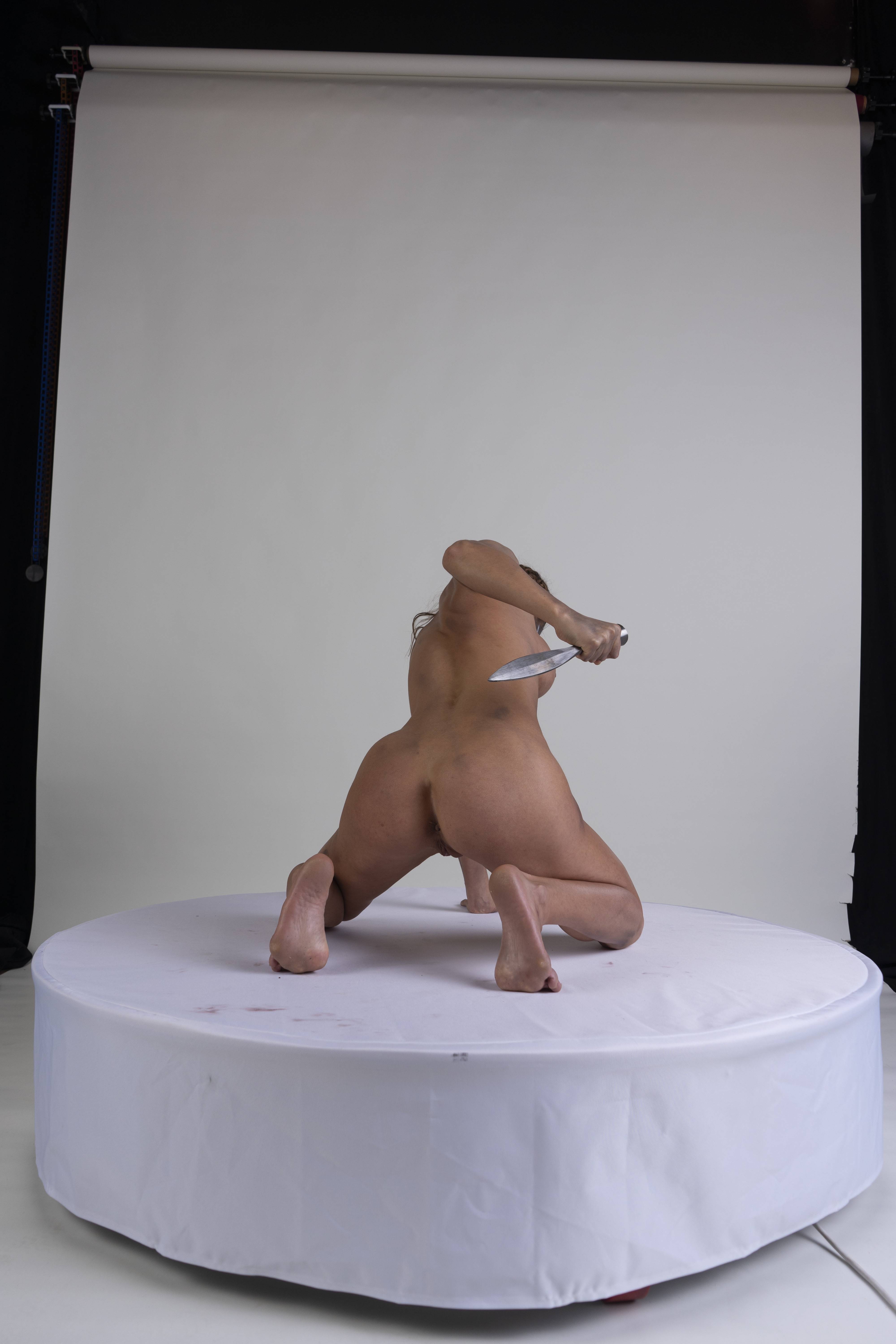 a nude female is crawling on a round white platform, facing away from the viewer. she is holding a knife behind her back. the background is a white studio backdrop.