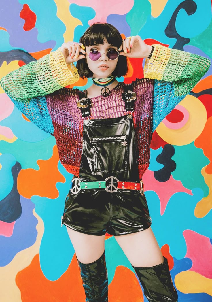 a woman is wearing a retroinspired outfit with a colorful psychedelic background. she is wearing sunglasses, a choker necklace with a peace sign, a rainbow crocheted sweater, shiny black overalls with a matching belt with peace sign buckles, and kneehigh boots. her hairstyle is short and bobbed with bangs.