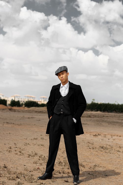 a full body shot of a man standing in a desertlike setting. he is wearing a black suit with a waistcoat, black overcoat, a newsboy cap, and black shoes. the sky is cloudy. he is looking directly at the camera with a serious expression.  the overall style is vintage or retro, reminiscent of the 1920s.