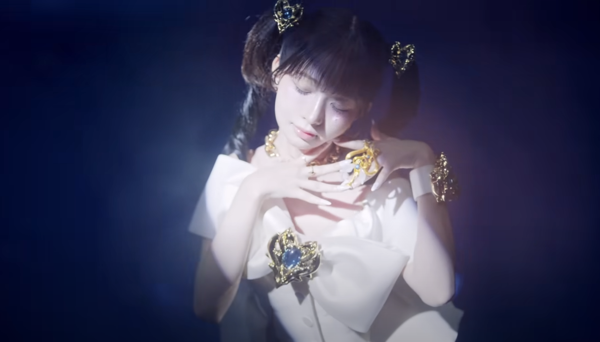 a young woman is the central focus of this image, likely a still from a music video or performance. she is heavily adorned with gold and blue jeweled accessories, contrasting against her white dress. she has her dark hair styled in two pigtails with decorative accents. the lighting is dramatic, a bright spotlight centered on her against a deep blue background. her pose and expression suggest a moment of intensity or allure within the performance.