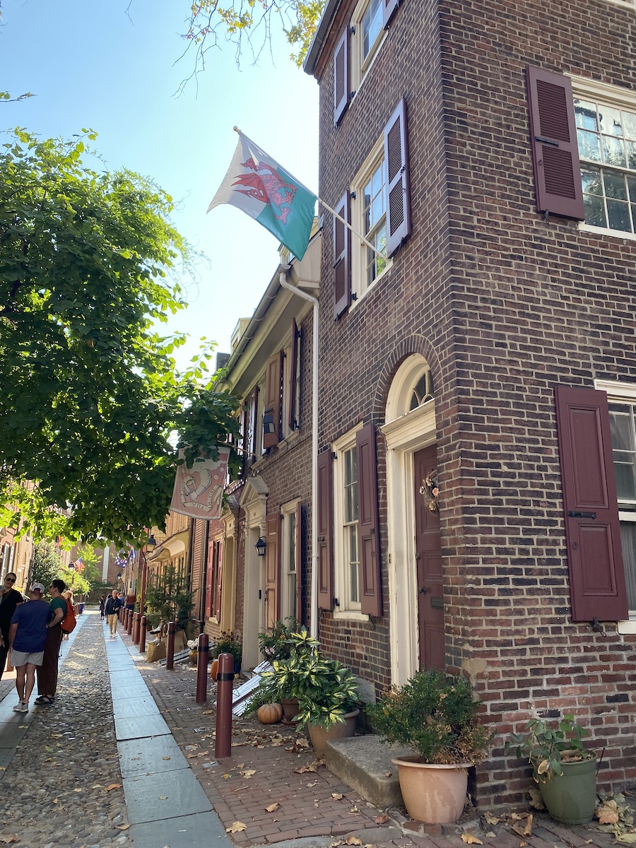 a view down a cobblestone street lined with brick buildings featuring shutters, doors, and windows, with a flag waving in the breeze and plants in planters along the sidewalk, along with people walking in the distance.