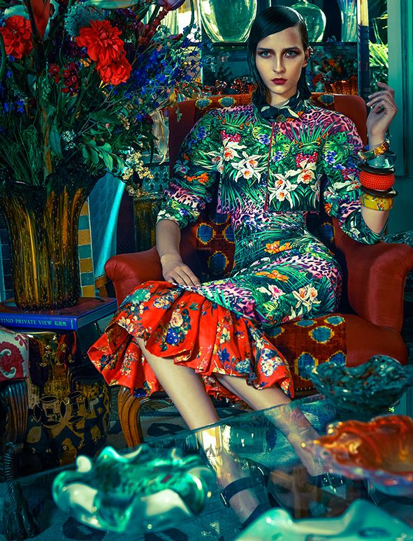 a fashion photograph showcases a stylish woman seated on a richly decorated armchair. she's wearing a vibrant, tropicalprinted dress with a ruffled hem, paired with striking jewelry. the surrounding interior is luxurious and eclectic, filled with colorful decor, artwork, flowers, and a vintage vibe. the overall mood is glamorous and sophisticated.