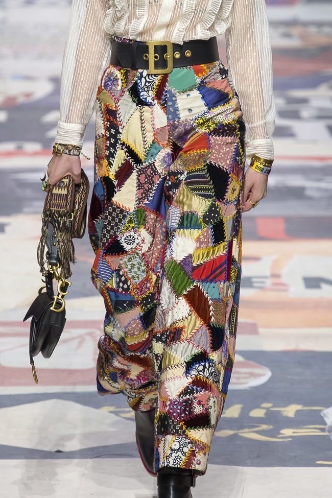 a person is shown wearing a white blouse with ruffle details, a wide black belt, and patchwork pants. the person is holding a black handbag and is wearing bracelets. the setting appears to be a runway.
