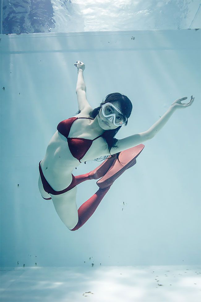 a young woman in a red bikini and diving mask is captured gracefully swimming underwater in a swimming pool. natural light streams through the surface of the water, illuminating her pose as she extends her arms and legs. the low angle shot emphasizes the feeling of weightlessness and freedom of movement underwater.