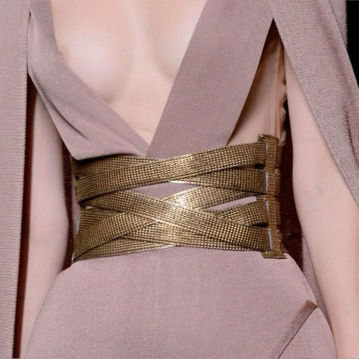 a closeup of a woman wearing a beautiful pink gown with a gold, metallic corset belt. the corset is made of delicate, woven bands and features a crisscross design. the model is posing on the runway, showcasing her elegant and glamorous style.