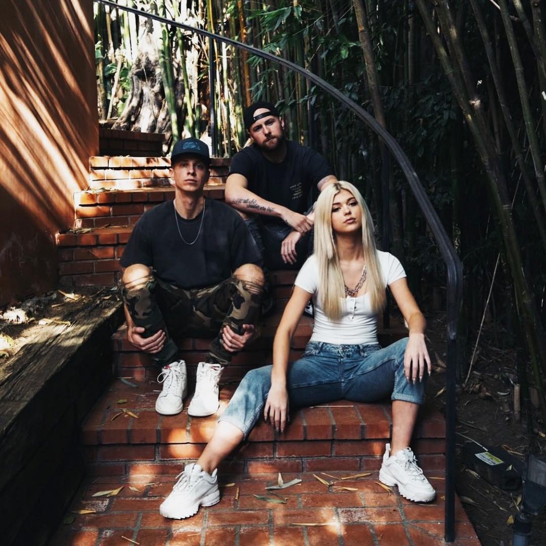 three people are sitting on brick stairs. two males and one female are in the image. the male on the left is wearing a dark tshirt, camo ripped jeans, white sneakers, and a baseball cap. he is also wearing a silver necklace. the male in the middle is wearing a dark tshirt, black baseball cap, and has tattoos on his arm. the female on the right is wearing a white tshirt, blue jeans, and white sneakers. there is a metal railing on the right side of the stairs. in the background there are trees and bamboo.