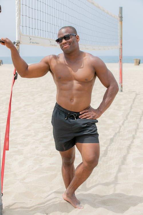 a muscular man with a tan, wearing black swim shorts and sunglasses, stands on a sandy beach leaning against a volleyball net. he is shirtless, showcasing his welldefined physique. the background shows the ocean and a clear sky. the overall mood is sunny and relaxed.