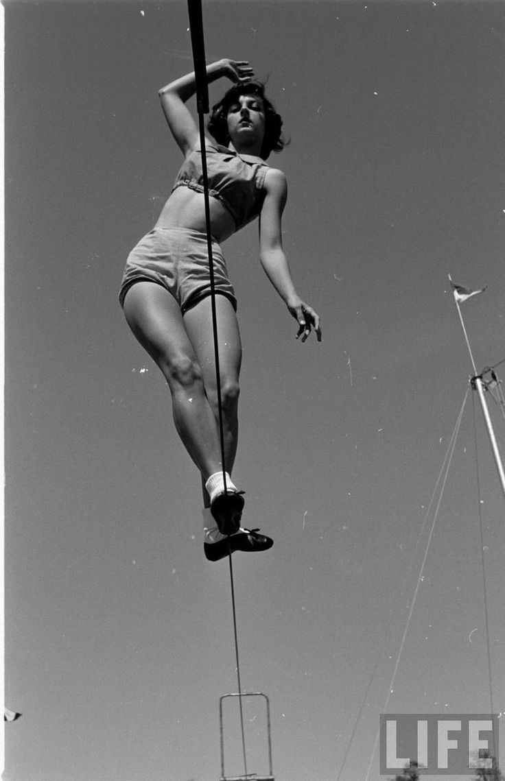 a black and white photo of a woman walking on a tightrope, taken outdoors. the woman is wearing a swimsuit and shoes. she is looking ahead. the photo is a vintage photo from life magazine.