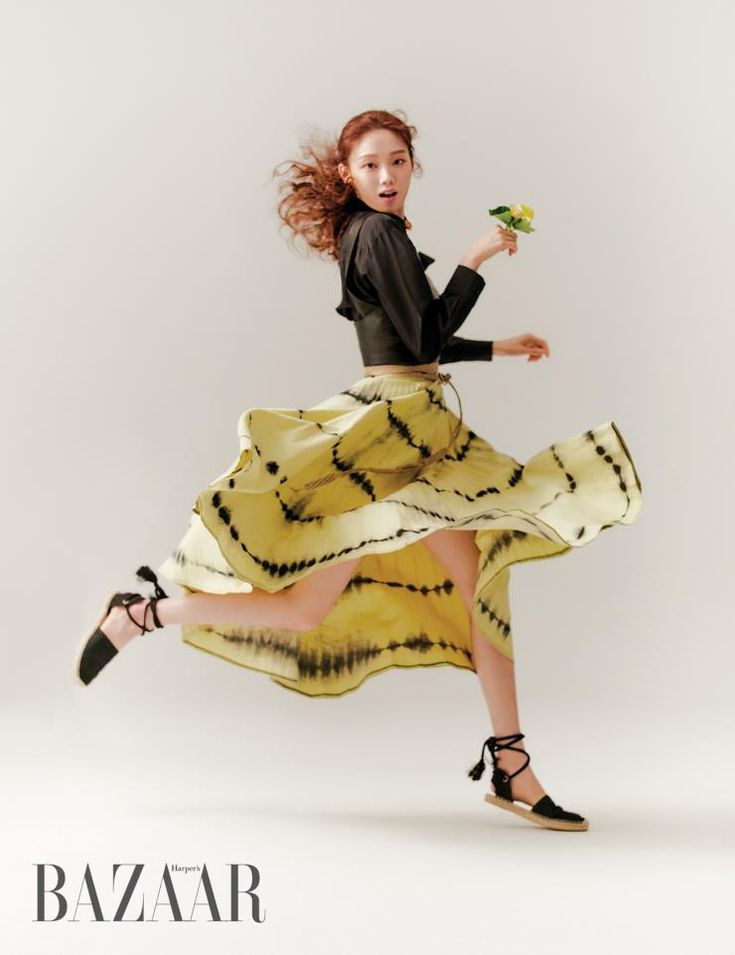 a young woman, wearing a black cropped shirt and a yellow and black tiedye skirt, is joyfully jumping and posing for the camera. she is smiling and has her hair in a curly style. she is holding a small bunch of flowers in her right hand. the background is a simple white studio background.  the image is captioned with harper's bazaar.