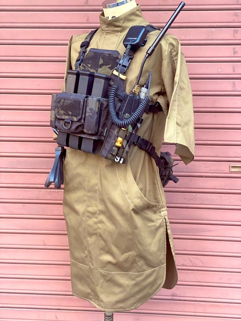 a mannequin wearing a tan colored outfit, with a tactical vest and gear,  is standing in front of a pink wall with a street in the background.