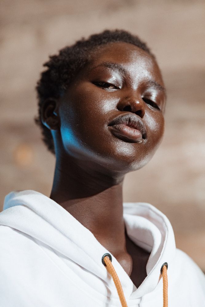 a closeup portrait of a young black woman looking up at the camera. she is wearing a white hoodie with an orange drawstring and has short hair. the woman is looking up slightly with a relaxed expression on her face.  her skin is glowing and there is a subtle shine to it, likely from makeup.  the background is out of focus, but it appears to be a brick wall.