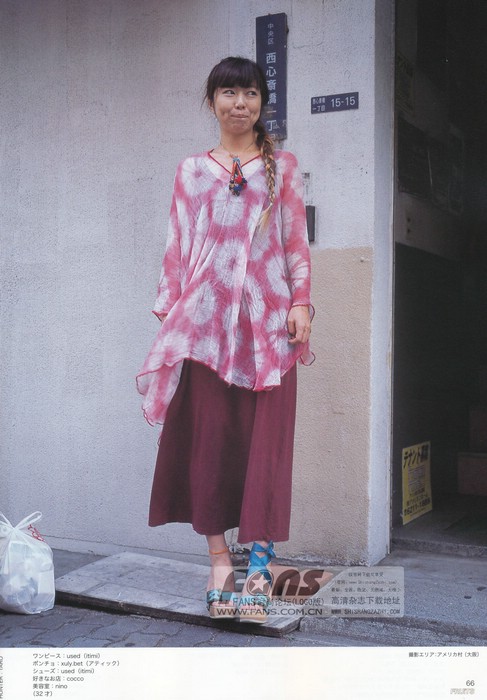 a young woman is standing on a sidewalk in front of a building. she is wearing a pink and white tiedye tunic top, a long maroon skirt, and brown sandals. she has long brown hair styled in a braid. she is carrying a white shopping bag in her left hand. the photo is taken from eye level and the lighting is natural daylight.