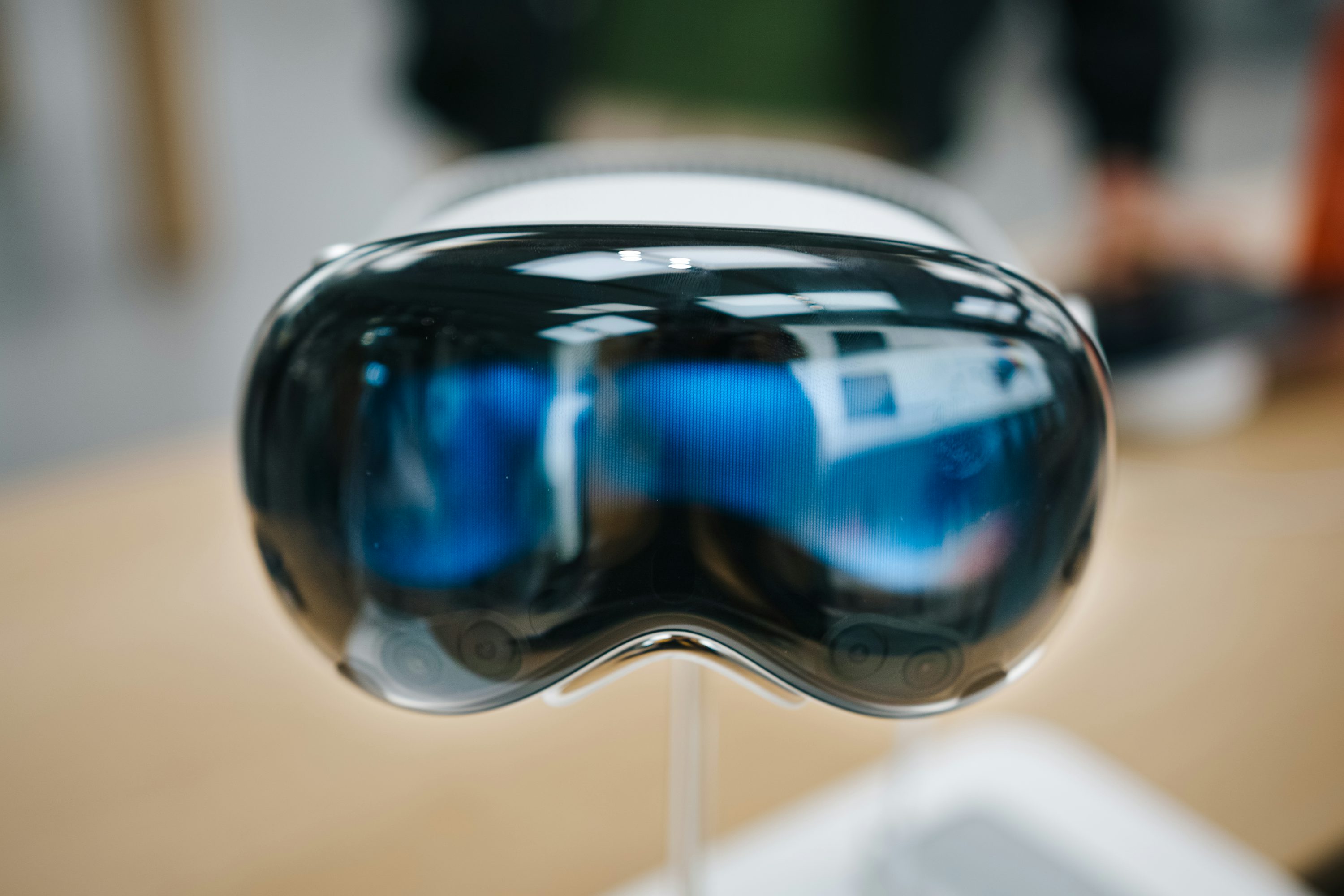 a closeup shot of the apple vision pro virtual reality headset. the headset has a glossy black front with bluetinted lenses, reflecting a digital interface. the device rests on a stand, with a blurred background suggesting it's in a retail or display setting.