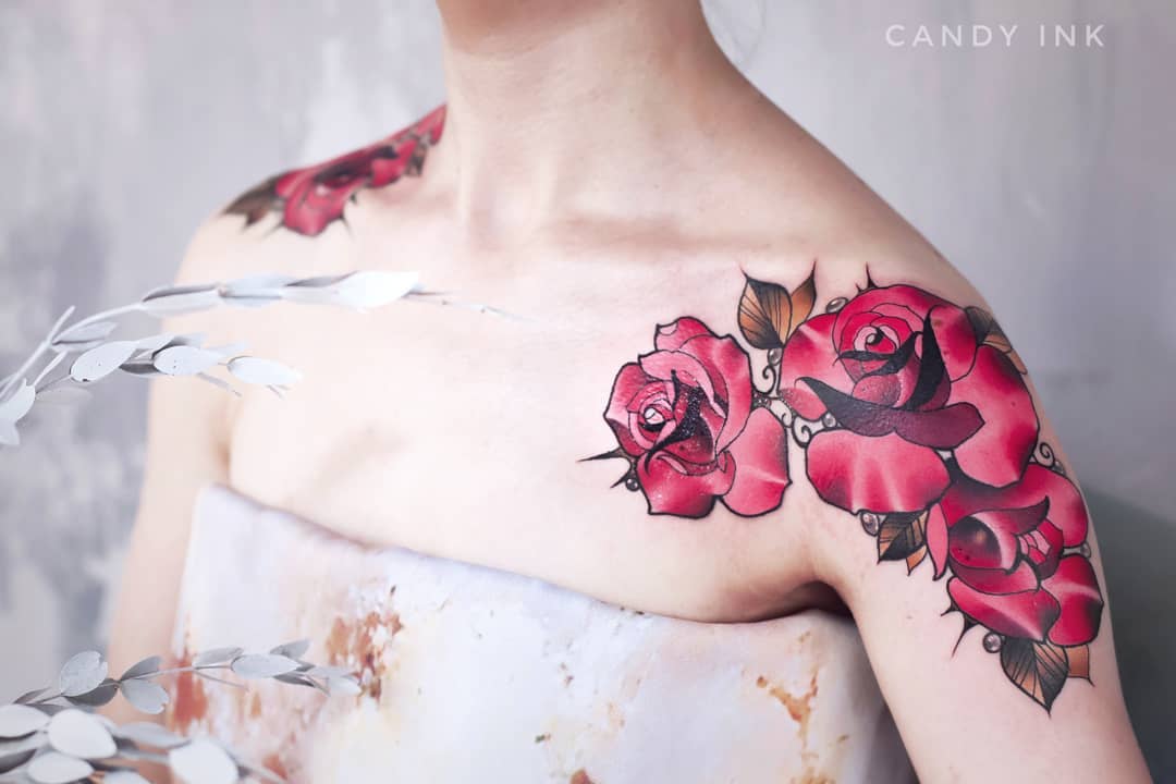 a closeup view of a woman's upper chest and shoulder showcasing a vibrant tattoo of red roses. the tattoo, done in a neotraditional style, features three prominent red roses with dark green leaves and subtle shading, creating a sense of depth. the woman's skin is fair, providing a stark contrast to the bold ink. the soft lighting and blurred background further accentuate the tattoo's intricate details.